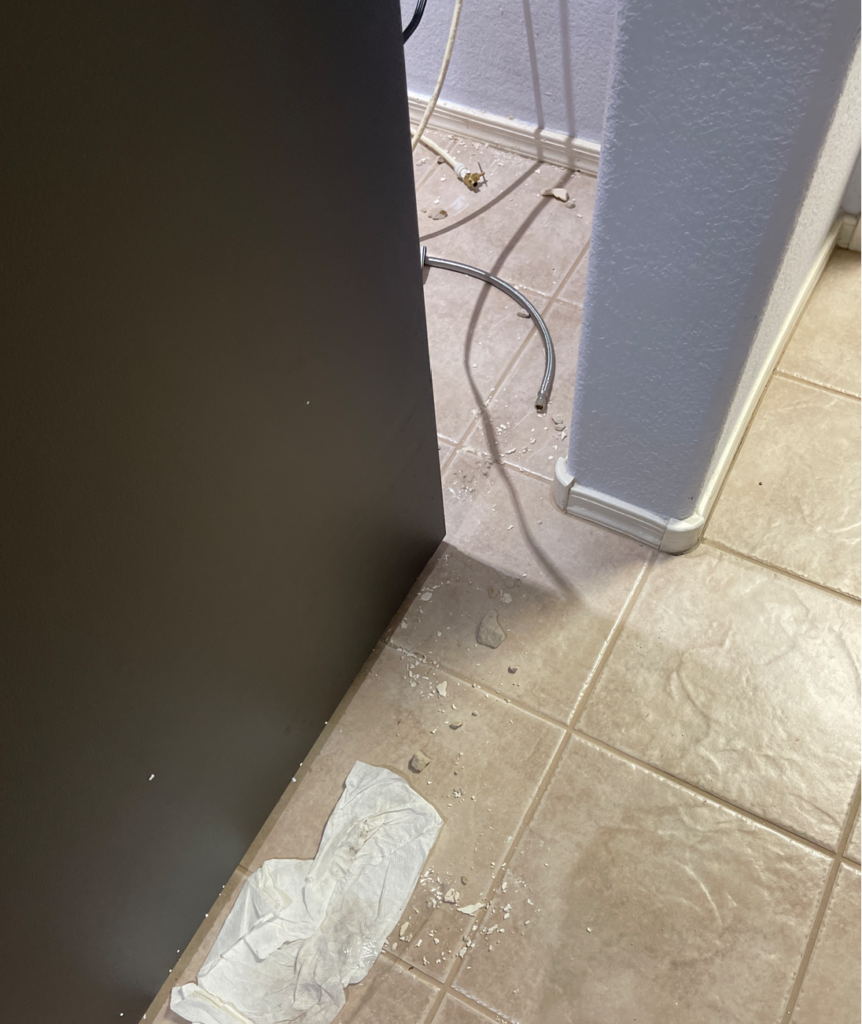 Some reminants of the debris left behind after the delivery team attempted to clean up. There are still chunks of drywall laying on the tile floor next to the new fridge, both water lines laying on the tile floor behind the fridge, and a used towel left next to the new fridge. 